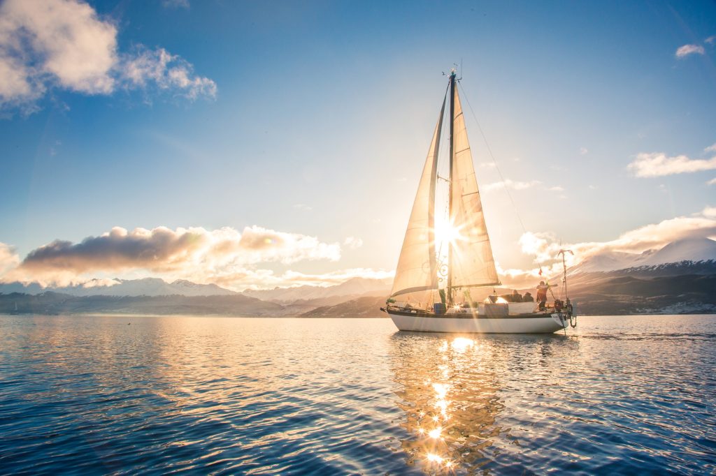 Un voilier naviguant paisiblement au coucher du soleil, sur une mer calme avec des montagnes en arrière-plan.